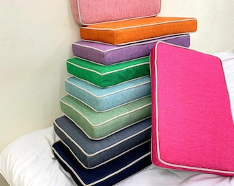 Puede incluir: Una pila de cojines rectangulares coloridos en varios tonos, incluyendo rosa, naranja, morado, verde y azul. Cada cojín tiene un ribete blanco. Los cojines están apilados uno encima del otro, con un cojín apoyado en la pila.