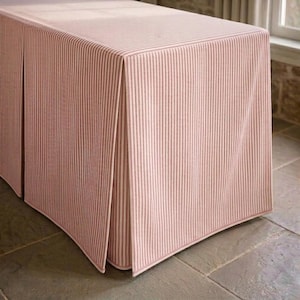 Puede incluir: Un mantel rectangular con un patrón de rayas verticales finas en rosa suave y blanco. La tela cae sobre los bordes de la mesa con pliegues estructurados y definidos en las esquinas, creando una apariencia entallada y uniforme.