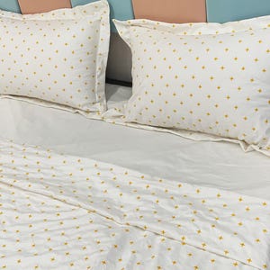 May include: White bed linen set featuring a repeating pattern of small yellow crosses. The set includes two pillowcases and a fitted sheet. The pillowcases have a ruffled edge. The bed is made up and ready for use.