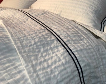 May include: White bedding with a quilted texture and black piping along the edges. The bedding is made of a soft, luxurious fabric.