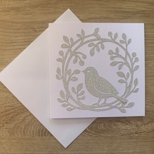 May include: A white card with a silver glitter design of a bird in a wreath of leaves.