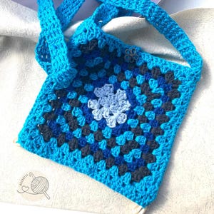 May include: A handmade crochet tote bag in shades of blue, with a floral design in the center. The bag features a square pattern and a strap for carrying. A small decorative flower is attached to the side.