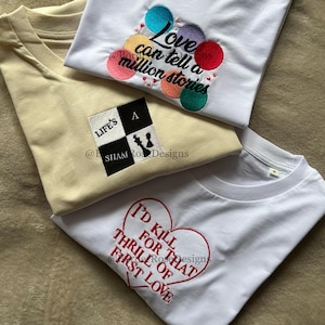 May include: Three t-shirts featuring embroidery. One white shirt displays colourful circles and the text "Love can tell a million stories". A beige shirt features a black and white square graphic with a chess piece and "LIFE'S A SHAM RO DESIGNS". A white shirt shows a red heart outline with the text "I'D KILL FOR THAT THRILL OF FIRST LOVE".