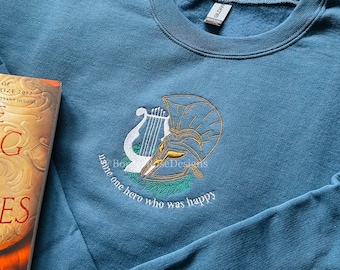 May include: A blue sweatshirt with a gold embroidered design of a lyre and a helmet. The text "Name one hero who was happy" is embroidered below the design.