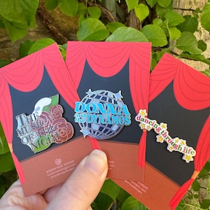 May include: Three enamel pins on display cards featuring red stage curtains. One pin shows a white mask and roses with the text The Music of the Night. Another is a blue disco ball with the text Donna and the Dynamos. The third says dancing through life.