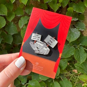 Peut inclure: Une épingle en émail argentée représentant une pile de journaux avec les mots "NEWSIES" imprimés sur le côté. L'épingle comprend également trois pancartes qui disent "STARE DOWN THE ODDS AND STRIKE!" et "SEIZE THE DAY!"