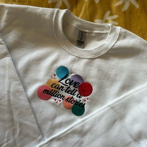 May include: White sweatshirt with a colorful embroidered design that says "Love can tell a million stories".