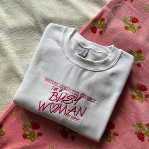 Pode incluir: Um moletom branco com o texto bordado "MATURE, COLLECTED, SENSIBLE, I'M A BUSY WOMAN (OH HEY)". O moletom est&aacute; sobre um cobertor rosa com estampa de morangos.