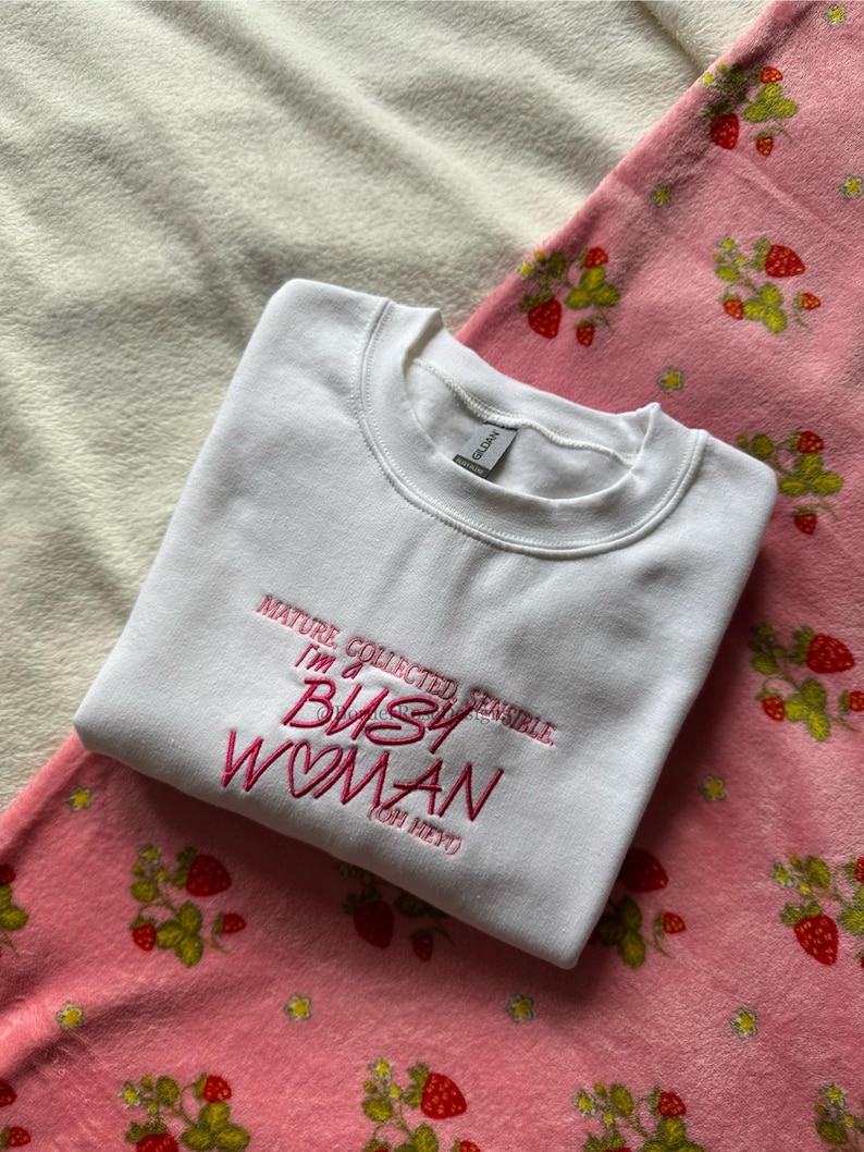 Pode incluir: Um moletom branco com o texto bordado "MATURE, COLLECTED, SENSIBLE, I'M A BUSY WOMAN (OH HEY)". O moletom est&aacute; sobre um cobertor rosa com estampa de morangos.