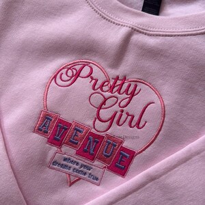 May include: A light pink sweatshirt with the embroidered text "Pretty Girl" inside a heart shape. Below the heart, the word "AVENUE" is embroidered on a series of angled blocks. The phrase "where your dreams come true" is also embroidered.