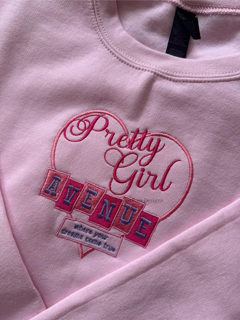 May include: A light pink sweatshirt with the embroidered text "Pretty Girl" inside a heart shape. Below the heart, the word "AVENUE" is embroidered on a series of angled blocks. The phrase "where your dreams come true" is also embroidered.
