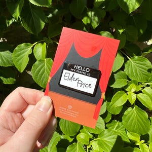 May include: A red and black card with the text "HELLO MY NAME IS" and the name "Elder Price" on a name tag. The card has a stage curtain design and the Border Rose Designs logo.