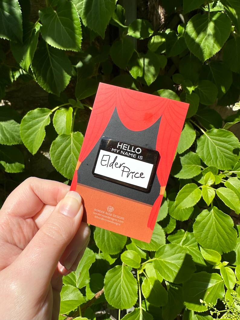 May include: A red and black card with the text "HELLO MY NAME IS" and the name "Elder Price" on a name tag. The card has a stage curtain design and the Border Rose Designs logo.