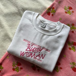 Pode incluir: Um moletom branco com a frase bordada em rosa: "MATURE, COLLECTED, SENSIBLE, I'M A BUSY WOMAN (OH HEY)". Est&aacute; sobre um cobertor rosa com estampa de morangos.