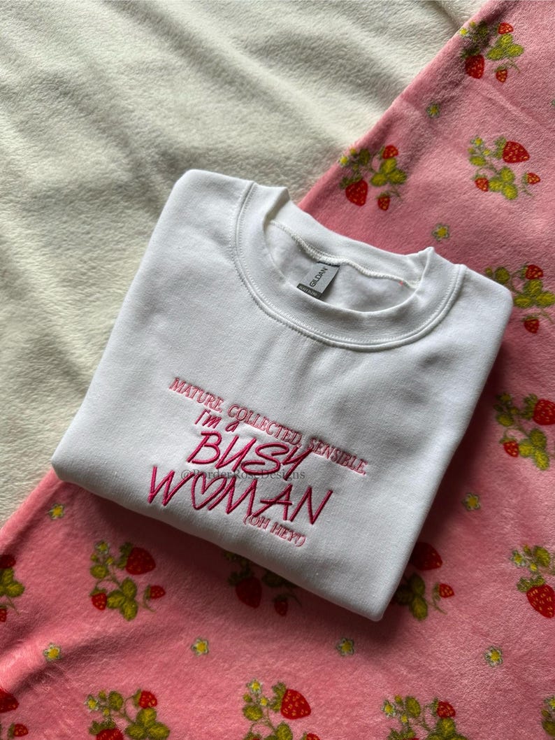 Pode incluir: Um moletom branco com a frase bordada em rosa: "MATURE, COLLECTED, SENSIBLE, I'M A BUSY WOMAN (OH HEY)". Est&aacute; sobre um cobertor rosa com estampa de morangos.
