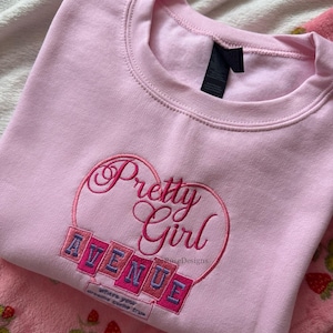 May include: A light pink sweatshirt with the embroidered words "Pretty Girl Avenue" in a heart-shaped design. The sweatshirt has a crew neck and a tag. The text "where your dreams come true" is also embroidered on the sweatshirt.