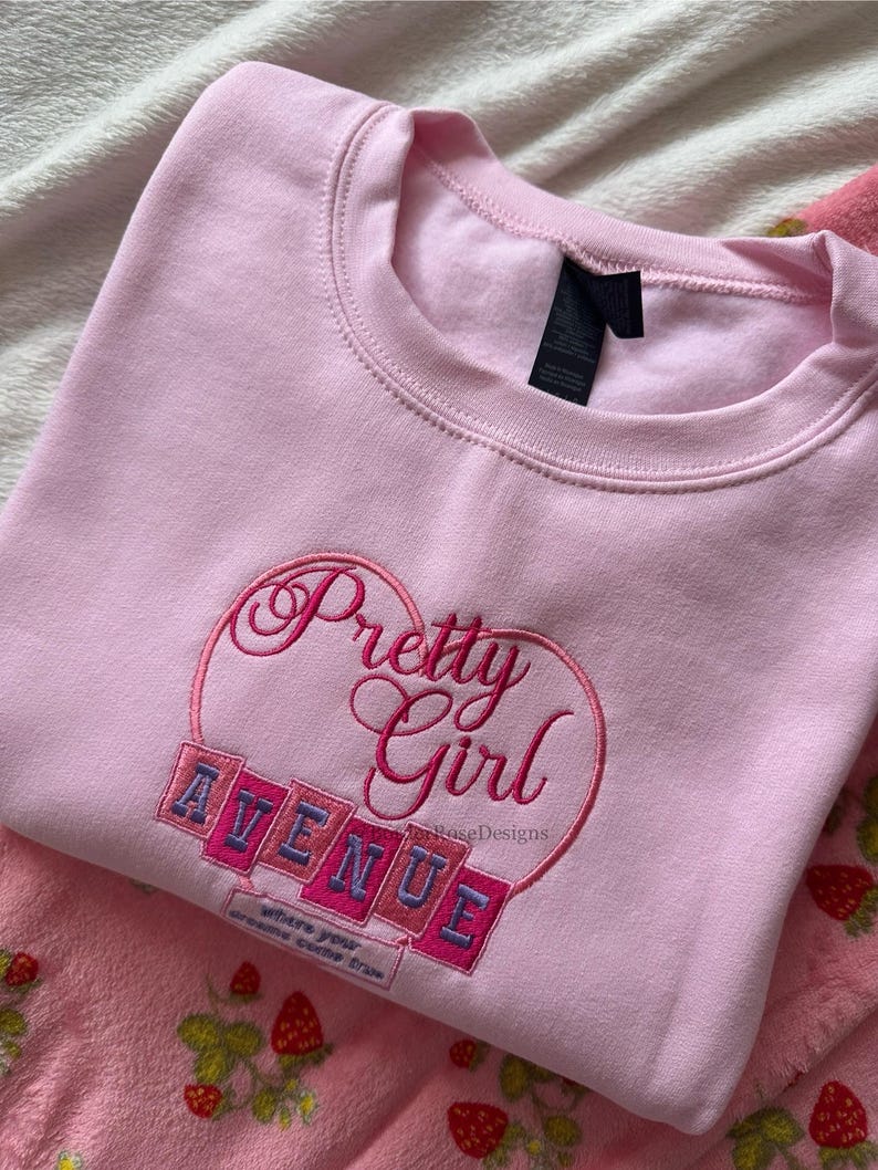 May include: A light pink sweatshirt with the embroidered words "Pretty Girl Avenue" in a heart-shaped design. The sweatshirt has a crew neck and a tag. The text "where your dreams come true" is also embroidered on the sweatshirt.