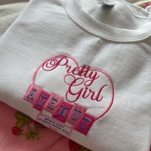 May include: White sweatshirt with the embroidered text "Pretty Girl Avenue" in pink and purple. The sweatshirt is folded on top of pink pajamas with a strawberry pattern. The phrase "where your dreams come true" is also embroidered.