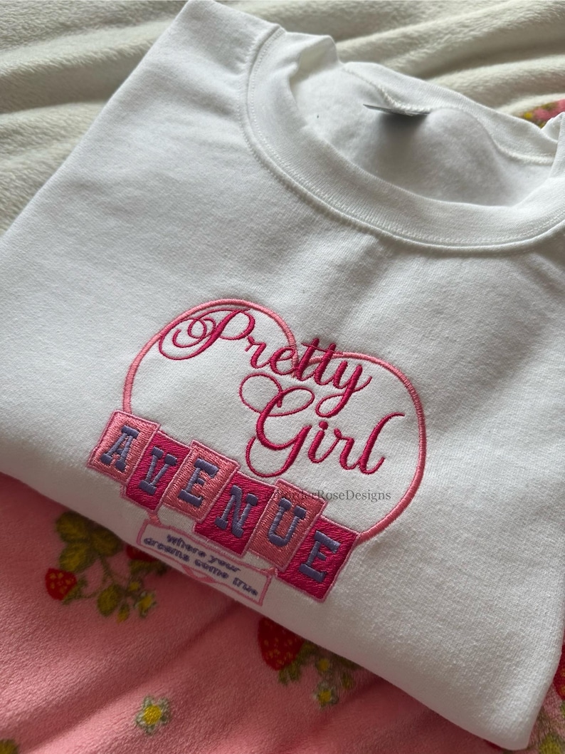 May include: White sweatshirt with the embroidered text "Pretty Girl Avenue" in pink and purple. The sweatshirt is folded on top of pink pajamas with a strawberry pattern. The phrase "where your dreams come true" is also embroidered.