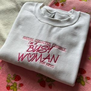 Pode incluir: Camisola branca de gola redonda com texto bordado em rosa: "MATURE, COLLECTED, SENSIBLE, I'm a BUSY WOMAN (OH HEY!)". A camisola est&aacute; dobrada sobre um cobertor rosa com um padr&atilde;o de morangos.