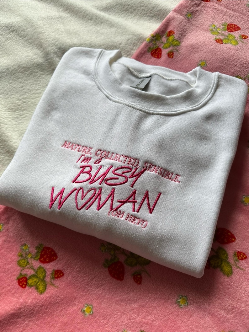 Pode incluir: Camisola branca de gola redonda com texto bordado em rosa: "MATURE, COLLECTED, SENSIBLE, I'm a BUSY WOMAN (OH HEY!)". A camisola est&aacute; dobrada sobre um cobertor rosa com um padr&atilde;o de morangos.