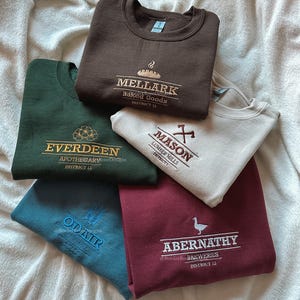 May include: Five folded sweatshirts in shades of brown, green, teal, cream, and burgundy. Each garment displays embroidered text and graphics, including "Mellark Baked Goods" and "Everdeen Apothecary". The sweatshirts are arranged on a white, textured surface.