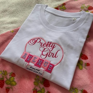 May include: A white t-shirt with the embroidered text "Pretty Girl Avenue" in pink and blue, set within a heart shape. The shirt is folded on a pink blanket with strawberry patterns.