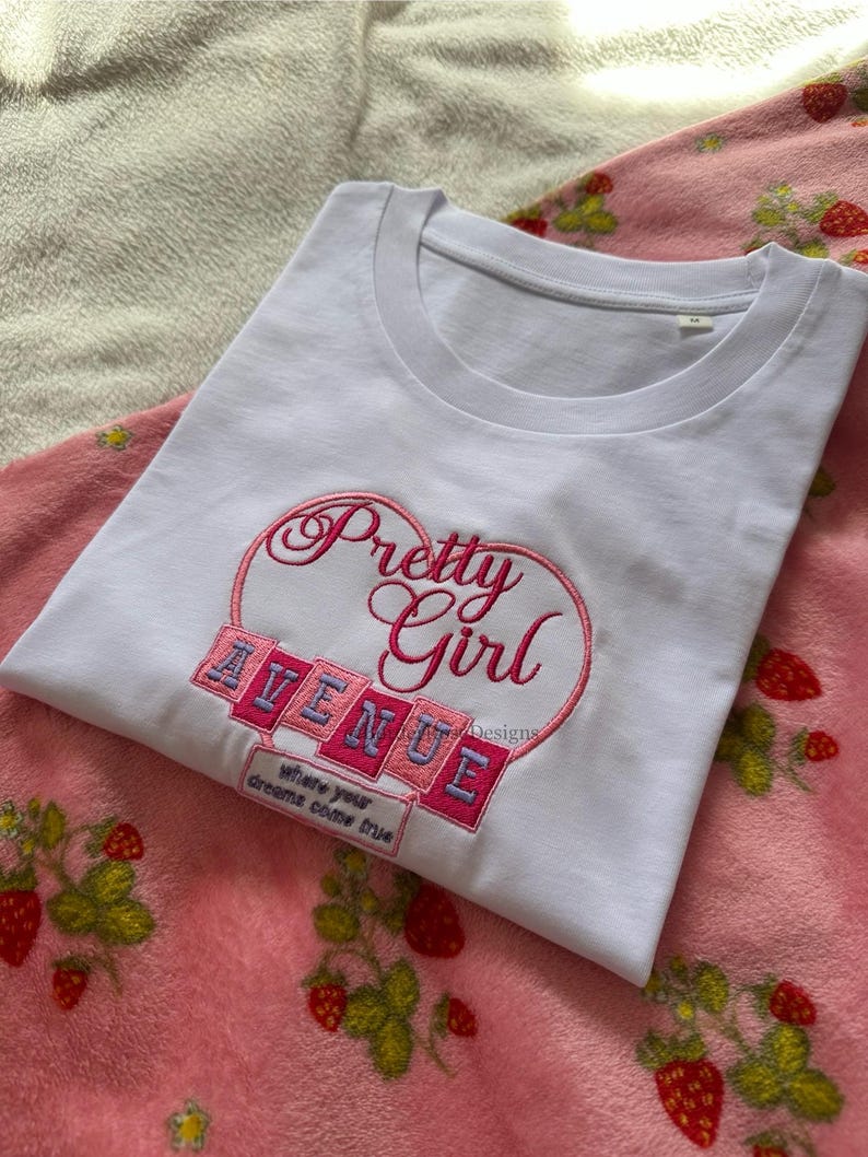 May include: A white t-shirt with the embroidered text "Pretty Girl Avenue" in pink and blue, set within a heart shape. The shirt is folded on a pink blanket with strawberry patterns.