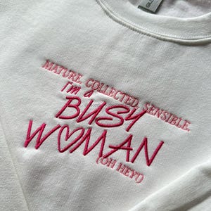Pode incluir: Camisola branca de gola redonda com texto bordado em rosa. O texto diz: "MATURE, COLLECTED, SENSIBLE. I'M A BUSY WOMAN (OH HEY)". A camisola tem uma etiqueta Gildan.