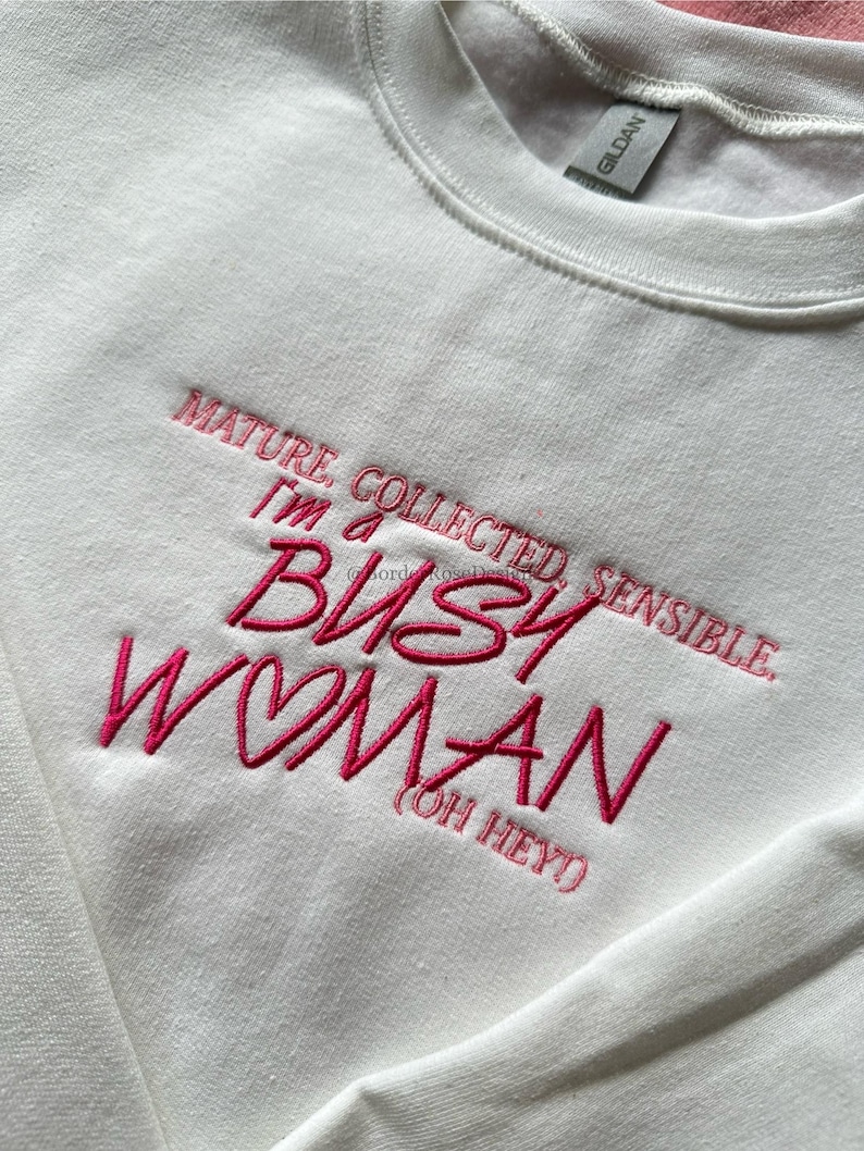 Pode incluir: Camisola branca de gola redonda com texto bordado em rosa. O texto diz: "MATURE, COLLECTED, SENSIBLE. I'M A BUSY WOMAN (OH HEY)". A camisola tem uma etiqueta Gildan.