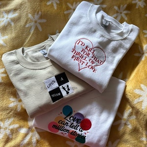 May include: Three white sweatshirts with embroidered designs. The top sweatshirt has a red heart with the text "I'd kill for that thrill of first love". The middle sweatshirt has a black and white chessboard with the text "Life's a sham". The bottom sweatshirt has colorful circles with the text "Love can tell a million stories".