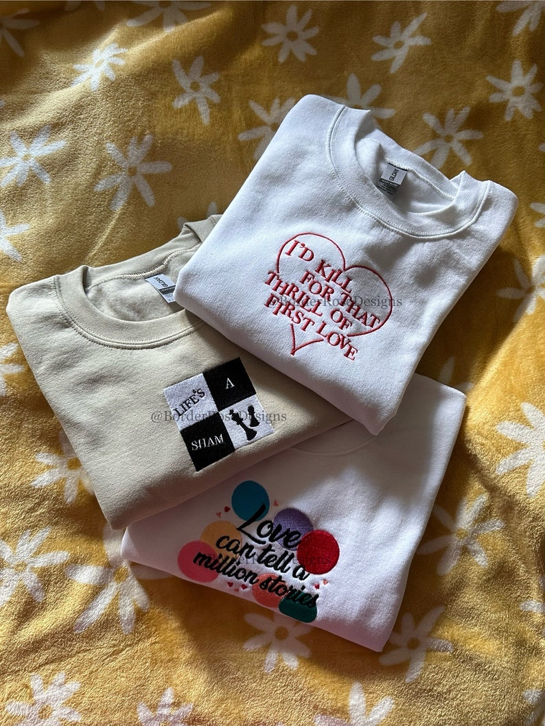 May include: Three white sweatshirts with embroidered designs. The top sweatshirt has a red heart with the text "I'd kill for that thrill of first love". The middle sweatshirt has a black and white chessboard with the text "Life's a sham". The bottom sweatshirt has colorful circles with the text "Love can tell a million stories".