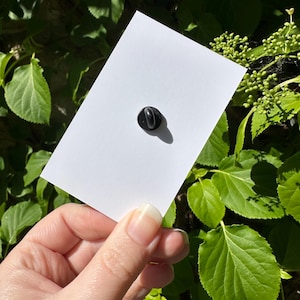 May include: A white rectangular enamel pin with a black pin back. The pin is held by a hand against a backdrop of green leaves and foliage. The pin is blank, ready for a custom design.