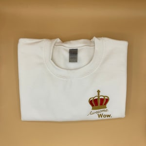 Puede incluir: Sudadera blanca con una corona bordada en oro y el texto "Awesome. Wow."