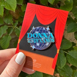 May include: A turquoise "DONNA and the DYNAMOS" pin set against a disco ball design, displayed on a card with a red curtain design. The card also features the brand name "BORDER ROSE DESIGNS" and its website address.