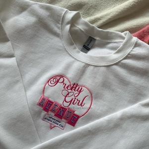 May include: White crewneck sweatshirt with a pink embroidered design. The design features the words "Pretty Girl Avenue" inside a heart shape, with the phrase "where your dreams come true" below. The sweatshirt is made of a soft material.