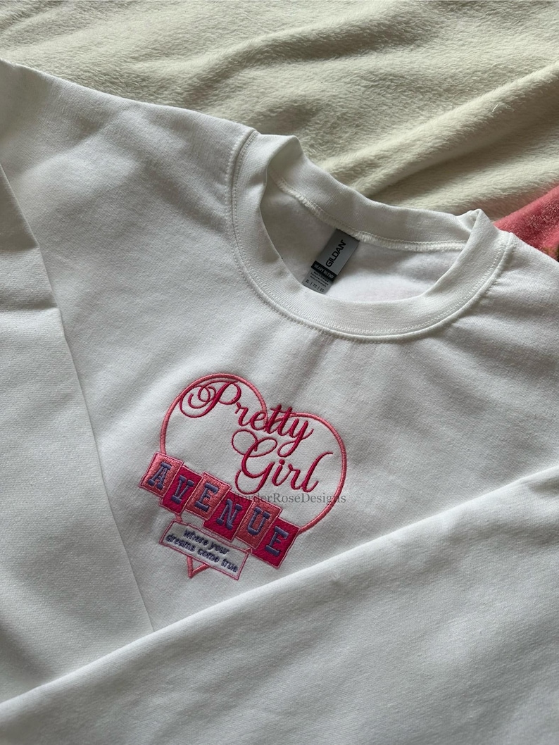 May include: White crewneck sweatshirt with a pink embroidered design. The design features the words "Pretty Girl Avenue" inside a heart shape, with the phrase "where your dreams come true" below. The sweatshirt is made of a soft material.