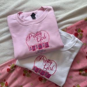 May include: Two sweatshirts, one pink and one white, with embroidered text. The pink sweatshirt features "Pretty Girl" in a heart design and "AVENUE" below. The white sweatshirt has the same design. Both are on a pink strawberry-patterned blanket.