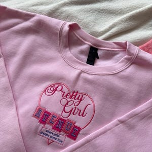 May include: A light pink sweatshirt with a crew neck. The front features an embroidered design with the words "Pretty Girl Avenue" inside a heart shape. Below the text is the phrase "where your dreams come true."