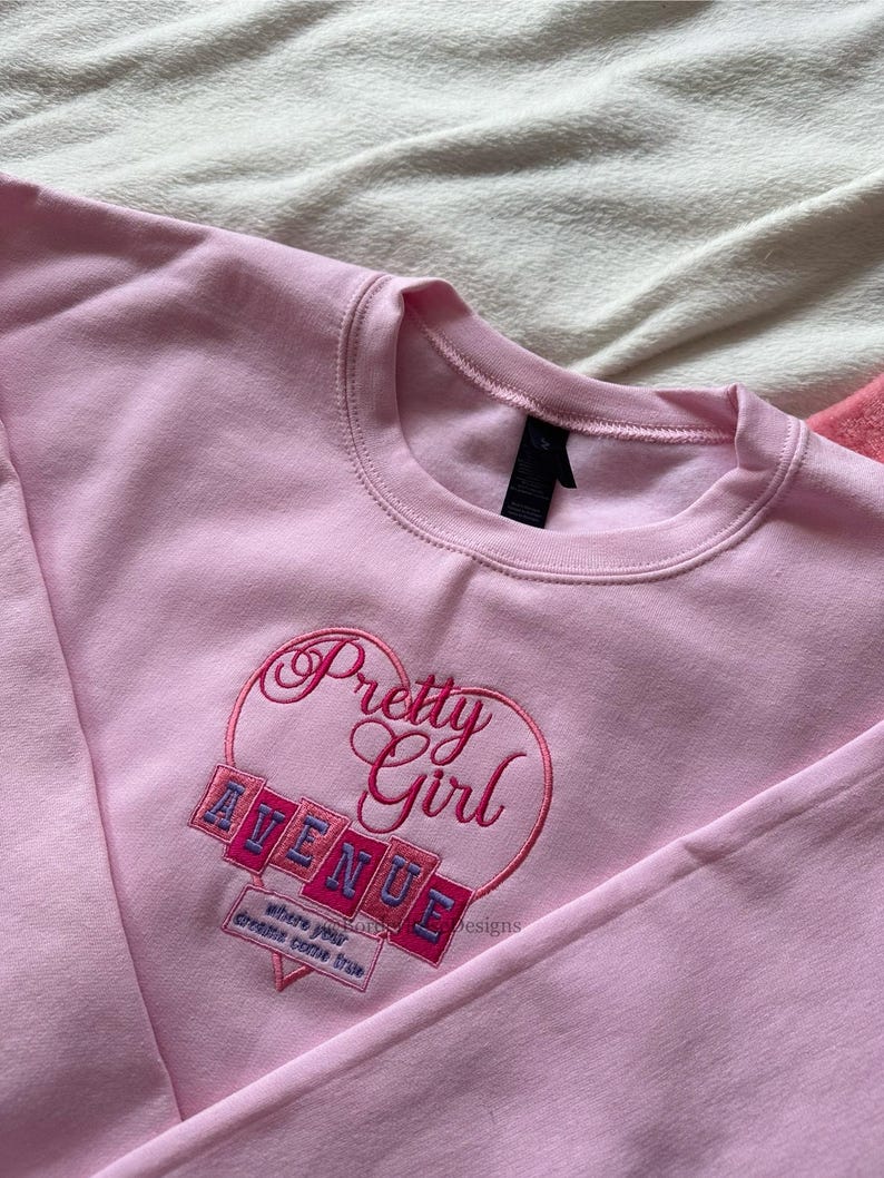 May include: A light pink sweatshirt with a crew neck. The front features an embroidered design with the words "Pretty Girl Avenue" inside a heart shape. Below the text is the phrase "where your dreams come true."