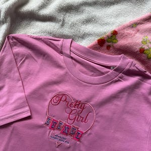 May include: A pink long-sleeve t-shirt with a heart-shaped embroidered design. The design includes the words "Pretty Girl" and "AVENUE" in a script font. The shirt is made of a soft material.