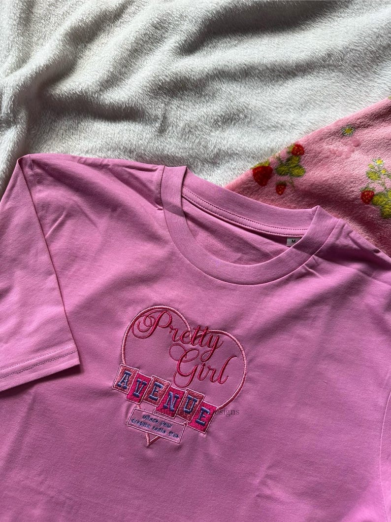 May include: A pink long-sleeve t-shirt with a heart-shaped embroidered design. The design includes the words "Pretty Girl" and "AVENUE" in a script font. The shirt is made of a soft material.