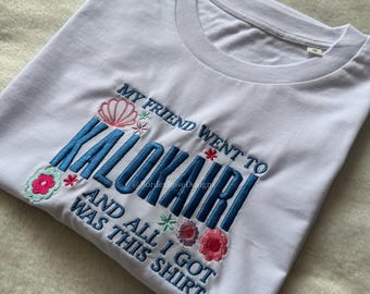 Może przedstawiać: Biała koszulka z niebieskim haftem: "MY FRIEND WENT TO KALOKAIRI AND ALL I GOT WAS THIS SHIRT." Tekst otoczony haftowanymi kwiatami w kolorze różowym, niebieskim i zielonym.