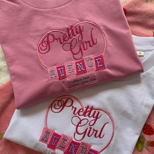 May include: Two t-shirts, one pink and one white, with embroidered designs. The pink shirt features a heart with "Pretty Girl" and "AVENUE" in a playful font. The white shirt has a similar design. Both shirts have the text "Where your dreams come true."