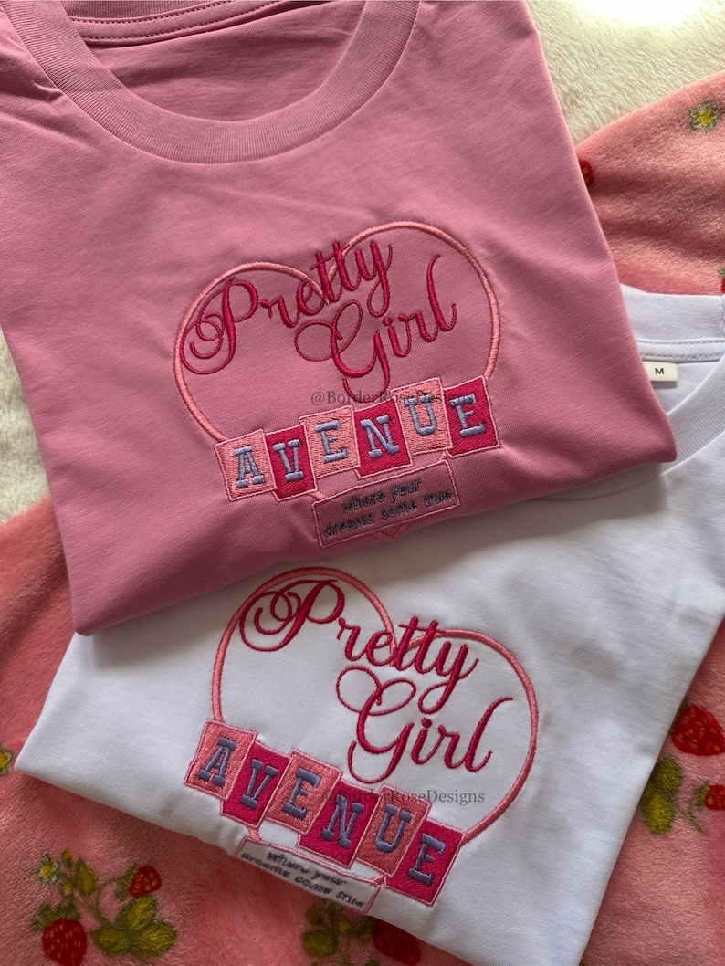 May include: Two t-shirts, one pink and one white, with embroidered designs. The pink shirt features a heart with "Pretty Girl" and "AVENUE" in a playful font. The white shirt has a similar design. Both shirts have the text "Where your dreams come true."