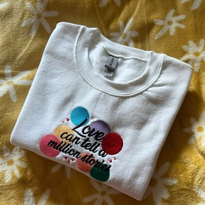 May include: White sweatshirt with a colorful embroidered design that says "Love can tell a million stories".
