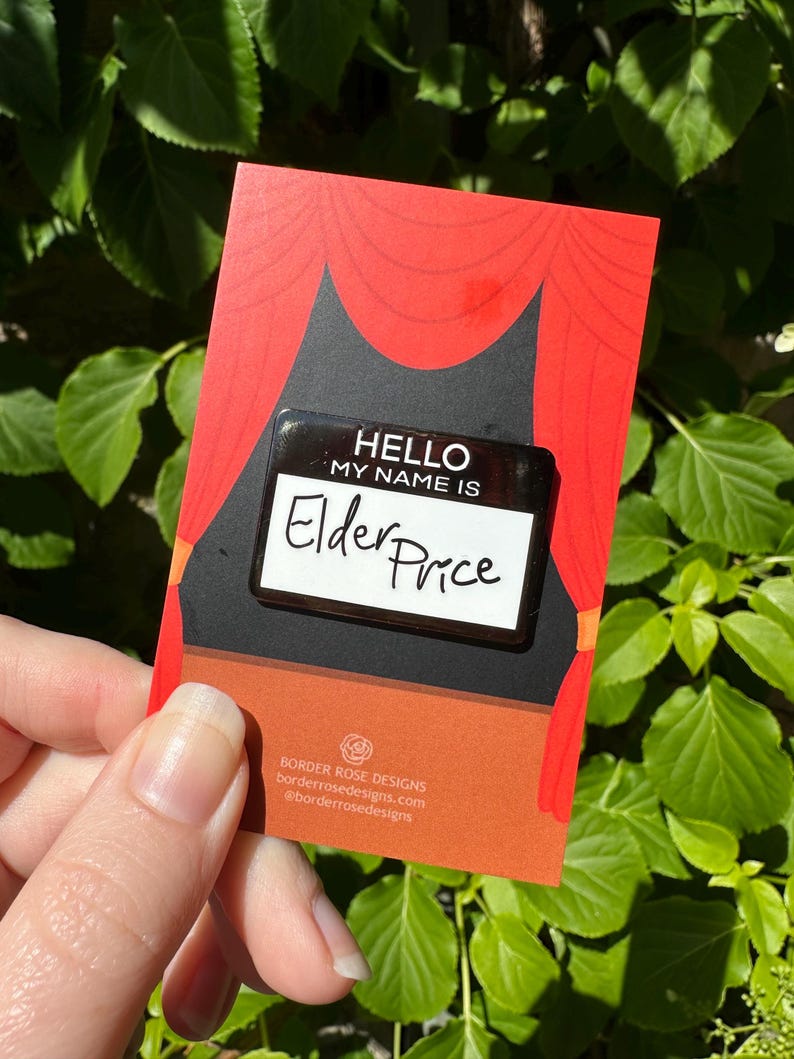 May include: A name tag pin with the text "HELLO MY NAME IS Elder Price" on a red and black card with a stage curtain design. The card also includes the Border Rose Designs logo and website address.