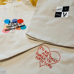 Peut inclure: Trois sacs fourre-tout en toile beige avec différents motifs. L'un affiche le texte "Love can tell a million stories" avec des cercles colorés. Un autre présente un motif sur le thème des échecs en noir et blanc. Le troisième sac a un cœur rouge avec le texte "I'd kill for that thrill of first love."