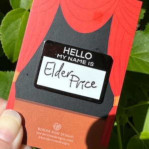 May include: A black and white name tag pin that reads "HELLO MY NAME IS Elder Price" on a red and black card with a stage curtain design. The card also includes the Border Rose Designs logo and website address.