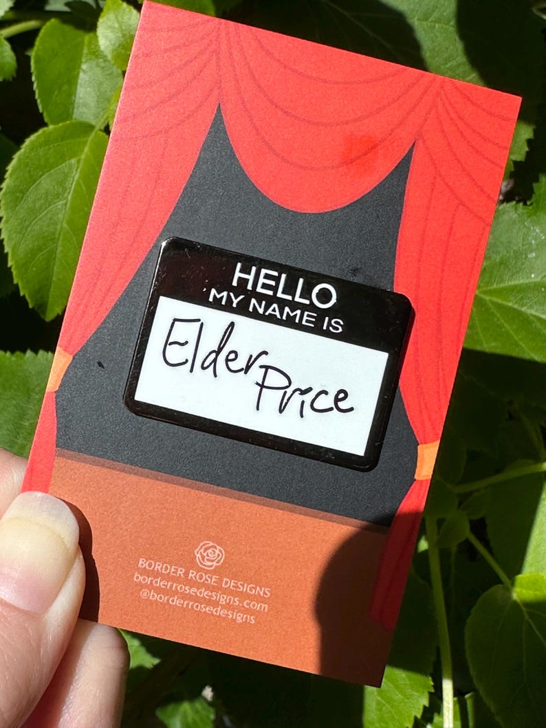 May include: A black and white name tag pin that reads "HELLO MY NAME IS Elder Price" on a red and black card with a stage curtain design. The card also includes the Border Rose Designs logo and website address.